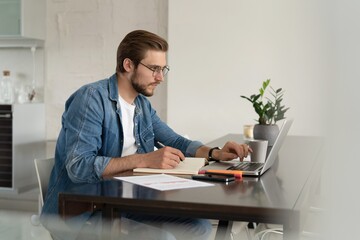 Young man working from home doing paperwork while using laptop.