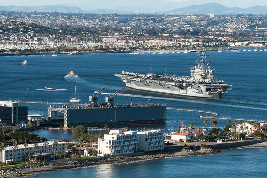 an aircraft carrier being led by a tugboat into San Diego Bay in California with a naval base in the foreground and the city of San Diego in the background