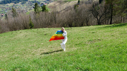 Young blonde woman with LGBT rainbow flag in the mountains on nature in white wedding dress.