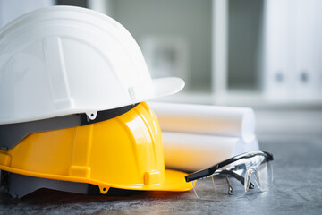 close up view of white and yellow hard safety helmet hat, blueprint, safety glasses on cement...