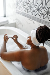 Rear view on shirtless naked overweight mixed race woman doing manicure inside of tub with foam, relaxing, doing beauty procedures. focus on female skin
