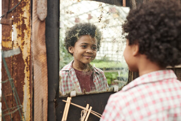 African little boy with curly hair looking at dirty mirror and smiling while standing outdoors