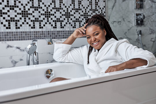 Adorable woman with perfect dark skin sits inside of bathtub, poses in bathroom enjoying weekends, self-care, romantic time alone