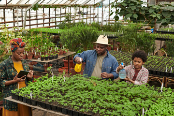 Family of three watering plants together while woman using digital tablet in the greenhouse