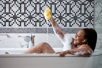 Smiling happy african woman clean fresh healthy black skin spa treatment enjoy relaxing taking shower and bath with bubble foam spa holding sponge in hands, in bathtub at bathroom at home - Powered by Adobe