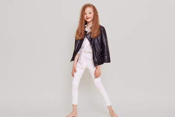 Full length portrait of confident schoolgirl wearing stylish attire and leather jacket, looking at camera, standing in trendy position, has blonde wavy hair, isolated over gray background.
