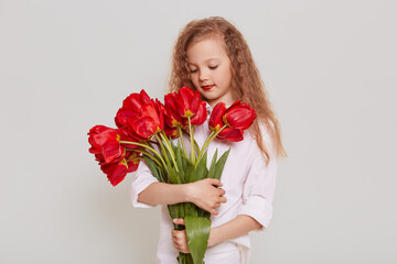 Charming blonde wavy haired girl wearing white blouse holding big bouquet of red tulips, looking at flowers with dreamy expression, isolated over gray background.