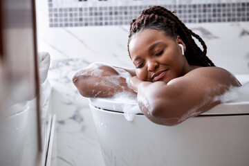Calm Dreamy Afro American Lady Lying In Bathtub With Foam, Relaxing In Headphones, With Eyes Closed, At Home In Bathroom