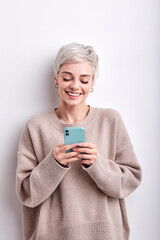 Portrait of pretty short haired woman in beige shirt chatting with friend using smartphone, typing message and smiling, looking at screen of mobile phone, isolated white studio background