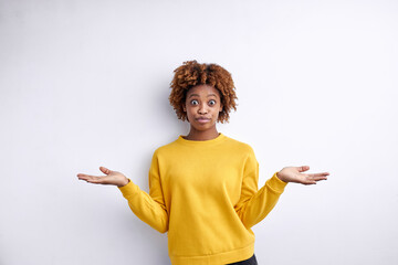 Dark skinned young woman with short curly haircut spreading arms and shrugging on white background, woman has doubts about choice or misunderstanding. Concept of human feelings.