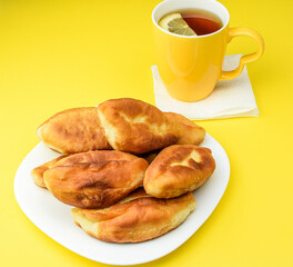 Lemon tea in a yellow ceramic mug and fried homemade pies on a white plate, on a yellow background. Tea party with pies. The concept of a hearty breakfast and snack. Top and side view.