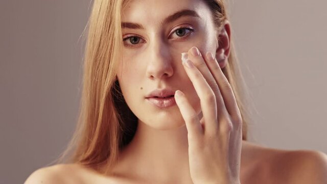 Female beauty. Silky facial care. Smooth lifting cosmetic. Moisturizing nutrition. Sensual lady tenderly applying cream on face cheek bone looking at camera isolated beige slow motion.