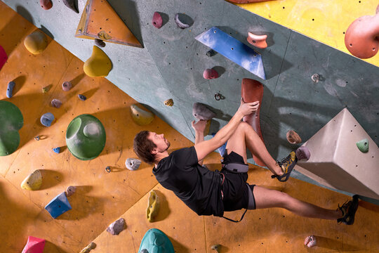 Free climber young man climbing on practical wall indoor, bouldering alone, in black sportive clothes outfit, professionally training. sport and workout concept