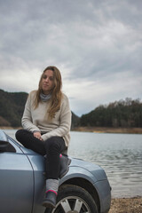 Young traveler girl sitting on top of the car in front of the lake