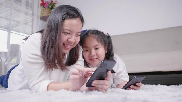 Asian mom and little daughter are lying on the floor in living room being on their phone together, mom enjoys .to show kid girl educational online program 
