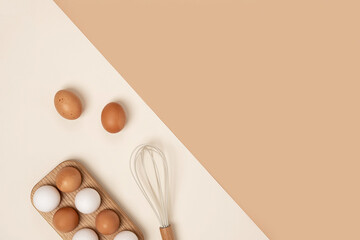 Eggs in wooden egg box and whisk on beige background. Flat lay Top view Copy space