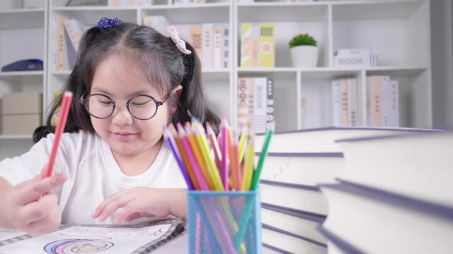 Asian girl wearing glasses is doing homework for school at the white deck, concentrating on her drawing and painting by coloured pencils in a cozy living room in her house after finishing school