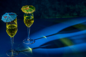 Two flute glasses with sparkling yellow liquid with colored paper umbrellas on top, cast reflections and shadows on a blue surface