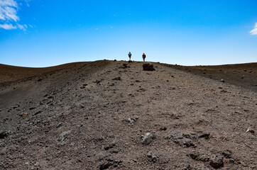 Two hikers walk on the crater rim of Etna volcano in Sicily, blue sky background 