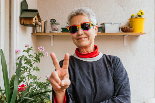 Lesbian older woman with short gray hair wearing rainbow pride flag sunglasses performing the victory sign on the terrace of her home. Human rights activist. lgbt community.