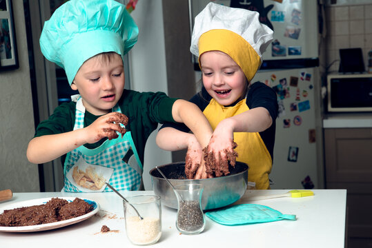 Siblings in cooking hats and aprons are buisy with kneading brown daugh in a bowl, both are happy, hands smudged. Concept of children cooking
