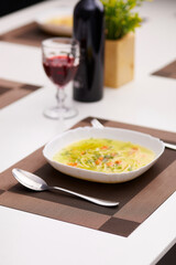 Subject photo of served dinner table with white plate of meal on rectangle brown and beige woven placemat. Spoon, fork, wine bottle, glass with ruby drink and pot flowers are located on the table. 