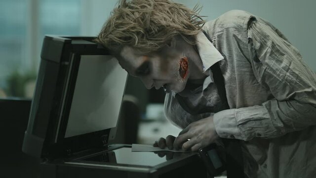 Medium shot of grunting zombie businessman in torn and dirty shirt and necktie and SFX makeup using copier in office