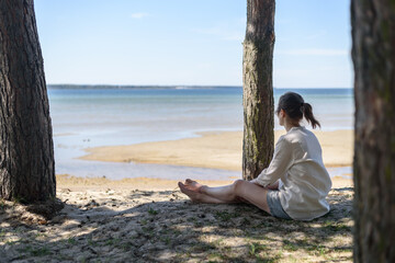 Woman in linen clothes sitting between pine trees and looking to sea. Wellbeing, mental health