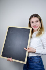 Portrait of a smiling beautiful young blonde woman holding an empty chalkboard vertically where you can add text and pointing it with the finger