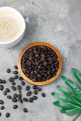 Biji Kopi or Coffee beans on wooden plate, with a cup of cappuccino and coffee beans on grey background. Top view. Copy space for text.