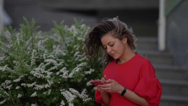 Portrait of a surprised woman in a red dress looking at a smartphone screen in the open air. Close-up of a girl reading a message on a mobile phone on a city street
