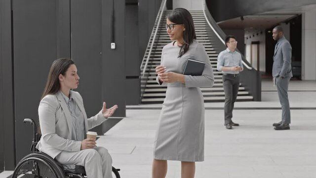 Lockdown of Caucasian handicapped female manager sitting in wheelchair, holding coffee paper cup while chatting with mixed-race female coworker standing in front of her