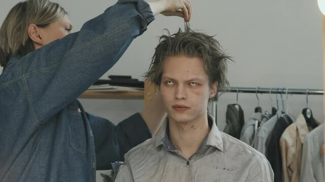 Handheld slowmo of female hairstylist backcombing hair of male horror movie or stage actor with scary contact lenses