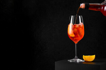 Pouring Aperol Spritz Cocktail in wine glass with ice and orange slice on dark background. Long fizzy drink