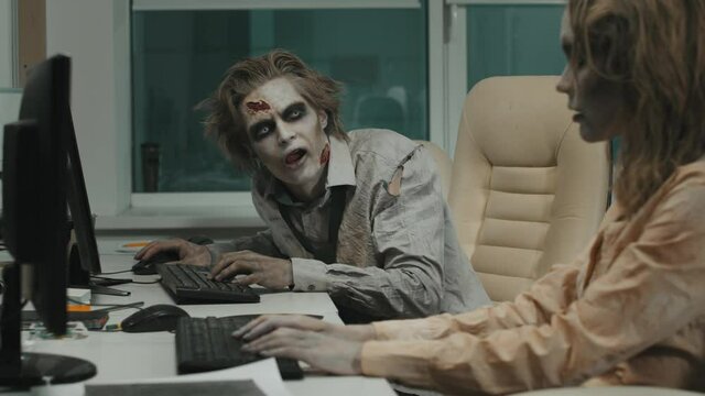 Medium shot with slowmo of mindless male and female zombies with SFX makeup and torn dirty clothes sitting at desks and smashing their hands on keyboard and mouse while trying to work on computers