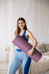 An attractive beautiful sportswoman smiles and stands with a gymnastic mat in her hands at home.