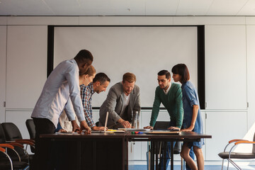 Group of six diverse business people bending over table in meeting room, discussing documents and...