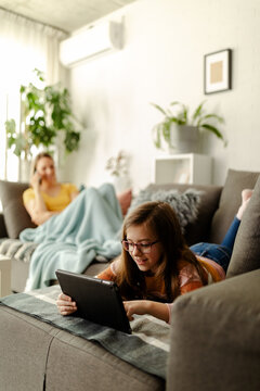 Little girl is laying on sofa at home and watching cartoons on tablet.