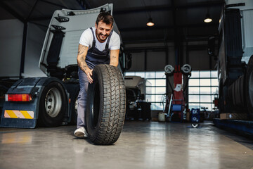 Smiling hardworking mechanic rolling tire in order to change it on truck. He is in garage of import...