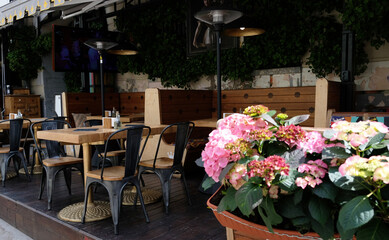 Fototapeta na wymiar Empty street patio restaurant, wooden table and chairs without people. Outdoor terrace of restaurant or summer street cafe. Colorful flowers hydrangeas in planters.