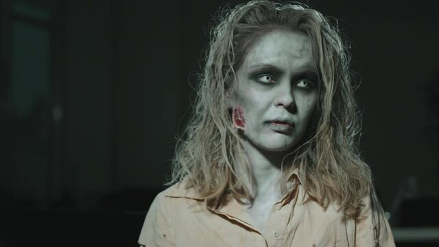 Medium shot of mindless female zombie with dirty hair, torn clothes and SFX makeup and contact lenses swaying and looking at camera in dark