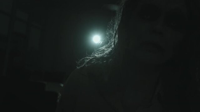 Close up shot of zombie woman with SFX makeup and contact lenses walking towards camera in darkness with spotlight shining in background
