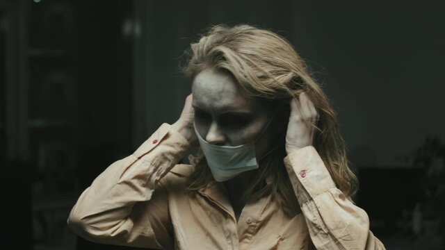 Portrait shot of zombie woman with SFX makeup and messy hair trying to put on face mask, then looking at camera in dark office