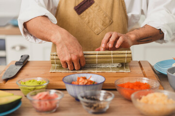Obraz na płótnie Canvas Asian chef hands rolling sushi with bamboo mat. Sushi preparation process with ingredients. Japan food delivery background
