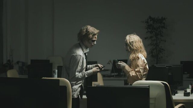 Medium shot of zombie man and woman holding mobile phones and bumping into each other while walking between desks in dark office