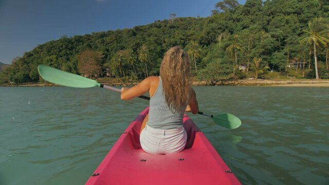 Long haired blonde woman with sunglasses rows bright pink canoe along sea bay water to beach with growing palms. Traveling to tropical countries. Sports girl is sailing on kayak in ocean.