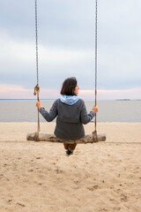 The girl sits alone on a large swing against the backdrop of a cloudy sunset sky.