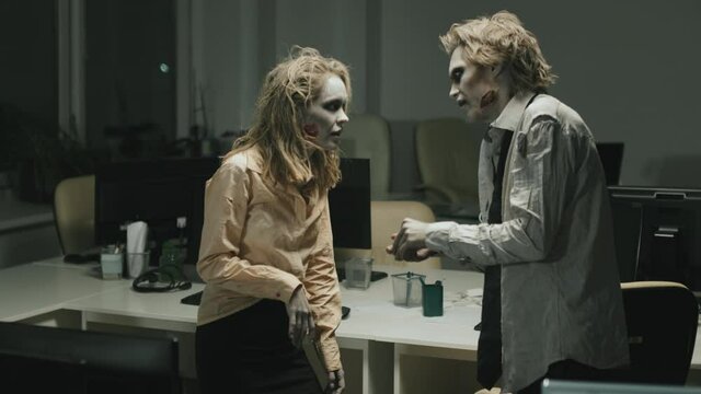 Slowmo medium shot of mindless female and male zombies with creepy makeup and dirty clothes trying to have conversation in office at night