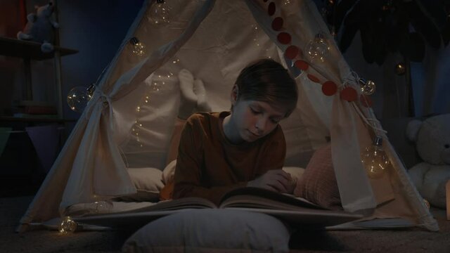 Teen boy in makeshift hut reading book at home in evening. Positive smart teenager lying on floor while spending free time. Concept of leisure and careless childhood.