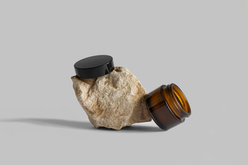 Opened brown glass jar with black cap laying, balancing on rock, gray background, copy space. Cream ,serum, oil, lotion on podium. Zero waste glass. Organic, natural cosmetic. Beauty, skincare product